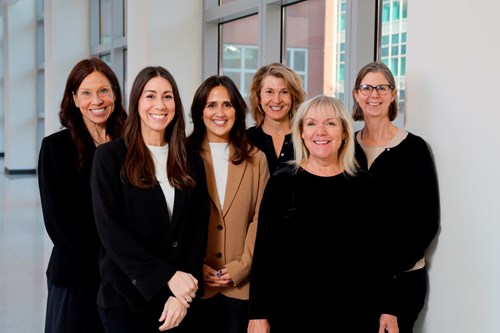 Six women in professional attire standing together in a bright indoor hallway near large windows